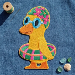 May include: An embroidered patch of a yellow duck wearing a pink floral swim cap, blue goggles, and a green and pink striped inner tube. The patch is placed on blue denim fabric next to a wooden spool of thread, a needle, and small sea shells.
