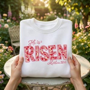 May include: White sweatshirt with the embroidered text "He is Risen" in pink, with the word "RISEN" filled with a pattern of red hearts. The text "Matthew 28:6" is also embroidered in pink.