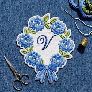May include: A circular embroidered patch featuring a white center with a dark blue cursive letter V, surrounded by a wreath of blue hydrangea flowers and green leaves, finished with a blue ribbon bow at the bottom, accompanied by scissors and thread.