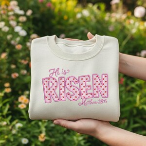 Peut inclure: Un sweat-shirt beige clair avec l'inscription "He is Risen" en rose, avec des cœurs. Le texte "Matthew 28:6" est en dessous. Le sweat-shirt est présenté sur un fond flou de feuillage vert et de fleurs.