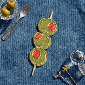 May include: An embroidered patch featuring three green olives with red pimentos threaded onto a silver cocktail pick. The patch is positioned on a blue denim fabric surface alongside a silver fork and a clear glass cocktail stirrer.
