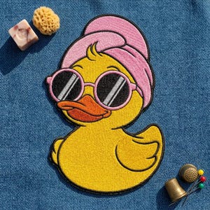 May include: An embroidered patch of a yellow rubber duck wearing pink round sunglasses and a pink towel wrapped around its head. The duck has an orange beak and is positioned on a blue denim fabric surface with a small sponge and a metal thimble.