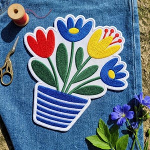 May include: A fabric patch featuring an embroidered bouquet of red, blue, and yellow flowers in a blue and white striped pot. The patch is placed on blue denim fabric next to a wooden spool of red thread and a pair of gold-toned metal scissors.