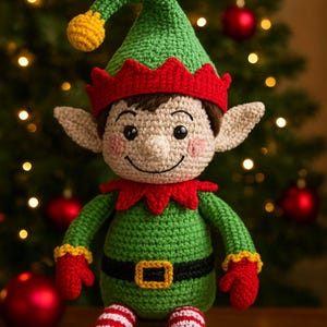 Könnte beinhalten: Gehäkelte Elfenpuppe mit grünem Hut und Outfit, rotem Kragen und Handschuhen sowie gestreiften Beinen. Der Elf hat ein lächelndes Gesicht, beige Haut und schwarze Schuhe. Im Hintergrund ein Weihnachtsbaum mit Lichtern und Ornamenten.