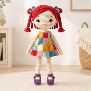 May include: A handmade crochet doll with red yarn hair styled in pigtails, adorned with blue bows. The doll wears a colorful patchwork dress and purple shoes. The doll is standing on a white rug.