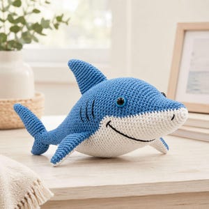 May include: A handmade blue and white crochet shark toy with black eyes and a smiling mouth. The shark has a dorsal fin, side fins, and a tail fin. The toy is sitting on a wooden surface.