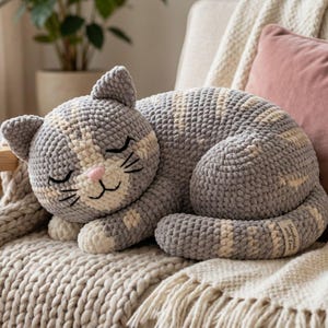 Peut inclure: Un coussin en forme de chat, fait à la main, de couleur grise et crème. Le chat endormi a les yeux fermés, un nez rose et des moustaches brodées noires. Le coussin repose sur une couverture crème.
