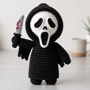 May include: A crocheted doll featuring a black hooded robe and a white mask with black eye and mouth openings. The figure holds a small grey knife with red stitched details in its right hand. The construction uses black and white yarn.