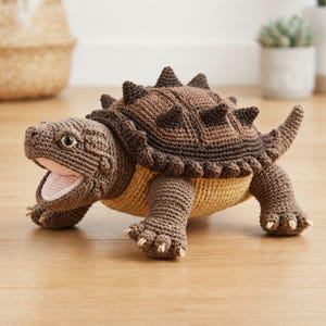 Puede incluir: Un juguete de tortuga caimán tejido a ganchillo con un caparazón texturizado de color marrón oscuro con púas prominentes, un vientre de color tostado claro y la boca abierta que revela un interior de color rosa pálido. Tiene cuatro patas con garras.