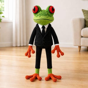 May include: A handmade crochet frog doll wearing a black suit, white shirt, and black tie. The frog has large red eyes and orange feet and hands. The frog's body is green, and the feet and hands are orange.