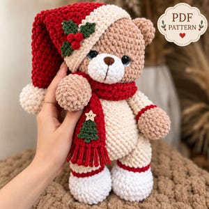 May include: A crocheted teddy bear wearing a red and white Santa hat with holly berries, a red scarf featuring a small green Christmas tree and a wooden star button, and white boots. The bear has light brown fur and a white muzzle. Text reads PDF PATTERN.