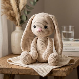 Puede incluir: Una muñeca de conejo beige tejida a ganchillo con largas orejas caídas, sentada sobre una alfombra cuadrada a juego. El conejo tiene ojos negros y una nariz rosa. La muñeca está sobre una mesa de madera, con un jarrón y libros al fondo.