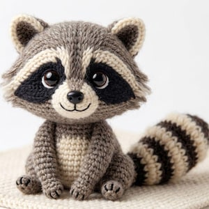 Puede incluir: Un juguete de mapache tejido a ganchillo con cuerpo gris, cara y pecho color crema y cola a rayas. El mapache tiene marcas negras en los ojos, una nariz negra y una boca sonriente. El juguete está sobre una superficie color crema.