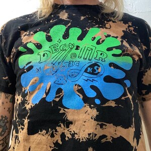 May include: Black t-shirt with a bleach-dye effect and a green and blue graphic design. The design includes the text "Decatur" and "KS" with other illegible text and illustrations. The shirt has short sleeves.