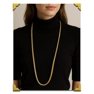 May include: A long, gold-colored chain necklace with a snake-like design. The necklace is worn over a black turtleneck sweater. The chain has a smooth, reflective surface and hangs down to the mid-torso.