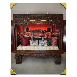 May include: A large, ornate wooden bed frame with intricate carvings. The bed is adorned with various pillows in shades of red, black, and patterned fabrics. The headboard features a red backdrop, and the bed frame sits on carved legs.