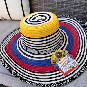 May include: A wide-brimmed hat with a colorful design. The hat features a yellow crown, a black and white striped band, and a brim with red, blue, and white stripes. A tag with text is attached to the hat.