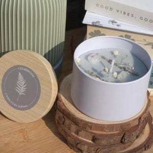 May include: A sage green ribbed ceramic vase next to a white candle tin containing light blue wax, dried flowers, and clear crystals. A round wooden lid with a grey label featuring a fern illustration and the text Cedarwood Candles by Sophia.