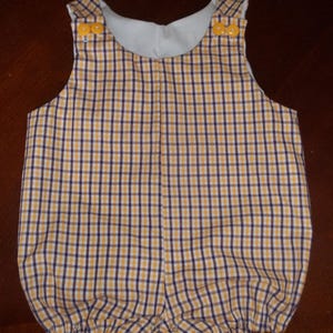 May include: A yellow, navy blue, and white plaid baby romper with yellow buttons on the shoulders. The romper has a rounded neckline and a gathered bottom. The fabric is a lightweight cotton, perfect for warm weather.
