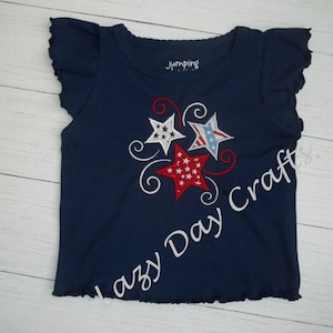 May include: A navy blue t-shirt with a white ruffle trim around the neckline and sleeves. The shirt features an embroidered design of three red, white, and blue stars with white swirls. The text "Jumping" is visible on the shirt.