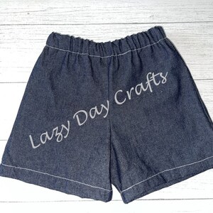 May include: A pair of denim shorts with a white elastic waistband and white stitching around the edges. The shorts have the text "Lazy Day Crafts" embroidered on the back in white.