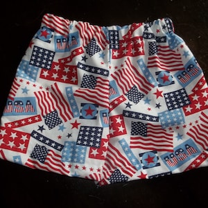 May include: Red, white, and blue patterned fabric shorts with a white background. The pattern features stars and American flags. The shorts have an elastic waistband.