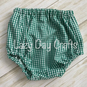 May include: Green and white gingham baby bloomers with a white elastic waistband. The bloomers are gathered at the legs and have a ruffled edge. The text "Lazy Day Crafts" is embroidered on the front of the bloomers.