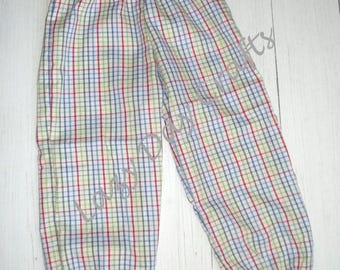 May include: A pair of pants featuring a multi-colored plaid pattern with thin red, blue, and green lines on a white background. The pants have an elastic waistband and elasticized cuffs at the bottom of each leg, providing a gathered appearance.