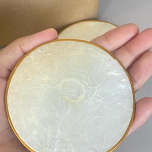 Puede incluir: Posavasos redondos, blanquecinos, con un borde dorado. Los posavasos tienen un aspecto nacarado y veteado. La imagen muestra dos posavasos, uno enfocado y otro ligeramente borroso en el fondo. Los posavasos están sostenidos en una mano.