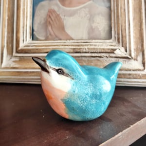 May include: A ceramic bird figurine in shades of turquoise, white, and peach. The bird has a black beak and eye details. The figurine is a decorative item, likely for display on a shelf or table.