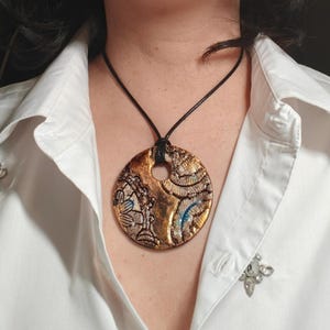 May include: A circular pendant featuring a textured, metallic bronze and gold finish with intricate embossed patterns and subtle blue accents. The pendant is suspended from a simple black cord, creating a bold contrast against the white collared shirt.
