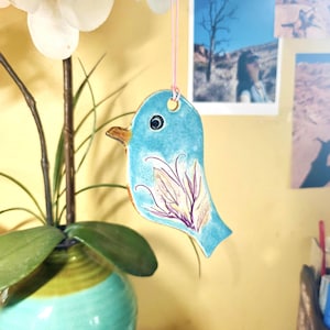May include: A light blue ceramic bird ornament hanging from a thin pink string. The bird features a dark eye, a brown beak, and a hand-painted floral design in shades of purple and cream on its side. The surface has a glossy, smooth finish.