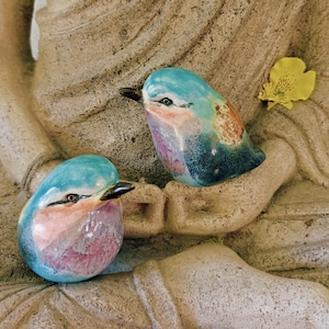 May include: Two smooth, rounded ceramic bird figurines featuring a glossy glaze in shades of turquoise, pink, and deep blue. Each bird has a small, dark, pointed beak and a painted eye, with one bird positioned slightly higher than the other.