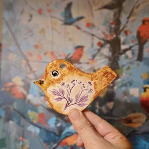 May include: A ceramic bird ornament with a glossy tan finish and a dark brown mottled texture. The bird features a small circular hole near the top for hanging and a detailed purple botanical illustration of a flowering plant on its side.