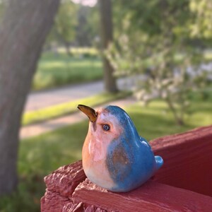 May include: A small ceramic bird figurine with a rounded body, featuring a glossy finish in shades of light blue and soft peach. The bird has a pointed brown beak, a single visible eye with a dark pupil, and a smooth, curved silhouette.