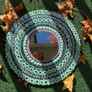May include: A round decorative mirror with a black frame, adorned with concentric circles of teal, blue, and purple embellishments. The mirror's surface reflects the sky. Scattered fall leaves surround the mirror.