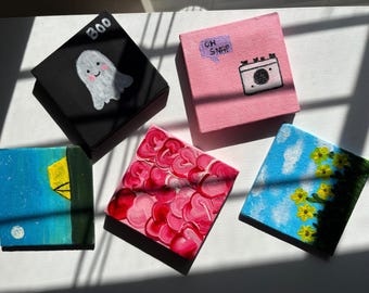 May include: Five small square paintings. A black canvas shows a white ghost with "Boo". A pink canvas has a camera and "Oh Snap". Other paintings depict flowers, a night scene, and abstract hearts. Each is a small artwork.