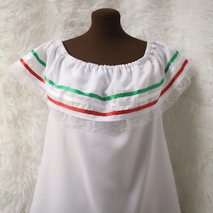 May include: White blouse with a scoop neckline, adorned with green and red ribbons, and lace trim. The blouse is displayed on a mannequin. The style is reminiscent of traditional Mexican attire.