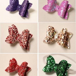May include: A collection of glittery hair bows in various colors, including purple, pink, red, gold, and green. The bows are arranged on a light-colored surface, showcasing their sparkly texture and decorative design. One bow is attached to a white elastic headband.