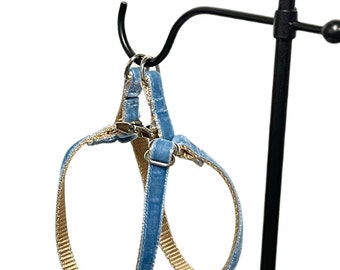 May include: A light blue velvet pet harness featuring a tan nylon webbing interior lining. The harness includes silver-toned metal hardware, including a central buckle and adjustable sliders, designed for securing around a pet's torso and neck.
