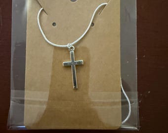 May include: A silver-colored cross pendant necklace displayed on a brown card inside a clear plastic bag. The cross is a simple, classic design, and the chain appears to be a thin snake chain. The packaging is simple and clean.