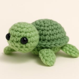 Può includere: Una piccola tartaruga all'uncinetto fatta a mano, nei toni del verde. La tartaruga ha una testa arrotondata, un guscio e quattro piccole pinne. È caratterizzata da due occhi neri di sicurezza. La tartaruga è un giocattolo carino e stravagante.