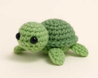 May include: A small, handmade crochet turtle in shades of green. The turtle has a rounded head, a shell, and four small flippers. It features two black safety eyes. The turtle is a cute, whimsical toy.