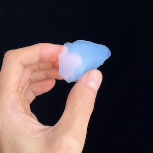 Puede incluir: Una piedra preciosa de ópalo en bruto, de color azul claro, con una superficie lisa y pulida. El ópalo se sostiene sobre un fondo negro, mostrando su cualidad translúcida y sutiles variaciones de color. La piedra tiene una forma natural e irregular.