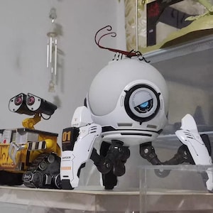 May include: A white spherical robot figure with three articulated legs, black accents, and a blue glowing eye, positioned next to a yellow and black tracked robot figure with two large circular eyes and the text WALL-E printed on its side.