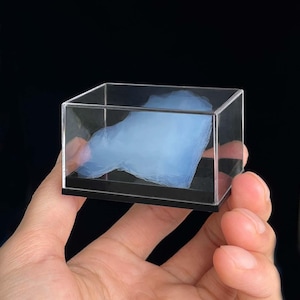 Puede incluir: Un objeto azul claro en forma de nube encerrado en una caja de acrílico transparente con una base negra. El objeto se sostiene en una mano sobre un fondo oscuro. El objeto en forma de nube parece estar hecho de un material suave y fibroso.
