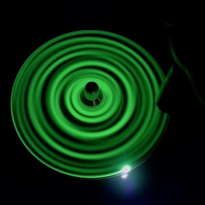May include: A spinning, glowing green object with concentric circular patterns against a black background. The object appears to be a disc or wheel, with a central hub. The motion creates a blurred effect, with bright green and dark green bands.
