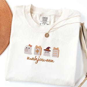 May include: A cream-colored t-shirt featuring an embroidered graphic of two ghosts, a mahjong tile, and a score card with the text "mahjoween" in orange script. The shirt has a Comfort Colors brand tag at the neck and a gold-toned pendant necklace.