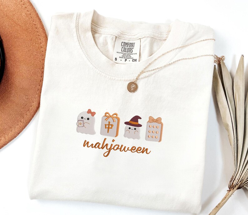May include: A cream-colored t-shirt featuring an embroidered graphic of two ghosts, a mahjong tile, and a score card with the text "mahjoween" in orange script. The shirt has a Comfort Colors brand tag at the neck and a gold-toned pendant necklace.