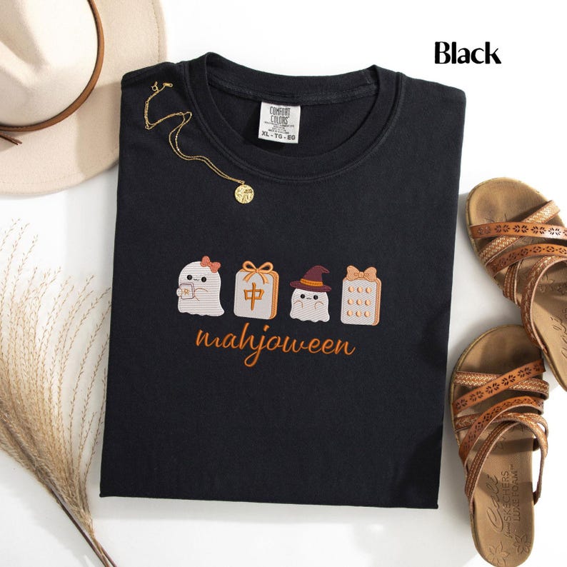 May include: A black t-shirt featuring a graphic of three ghosts and a mahjong tile with the text "mahjoween" printed below. The first ghost wears a bow, the second is a mahjong tile, and the third wears a witch hat. A gold necklace rests on the shirt.